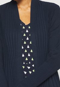 Cardigan blu navy lavorato a maglia con un design a coste testurizzato. Indossato sopra un abito scuro con un motivo di pere bianche e verdi.