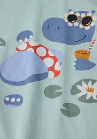 Vaaleansininen kangas, jossa on sarjakuvamainen hippo aurinkolaseissa, punaisissa pilkuissa ja juoma, ympärillä koristekiviä ja lumpeita.