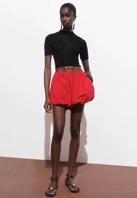 Zwarte geribbelde crop top gecombineerd met rode volumineuze shorts. Model staat op een bruine ondergrond met donkere sandalen en grote ringoorbellen.
