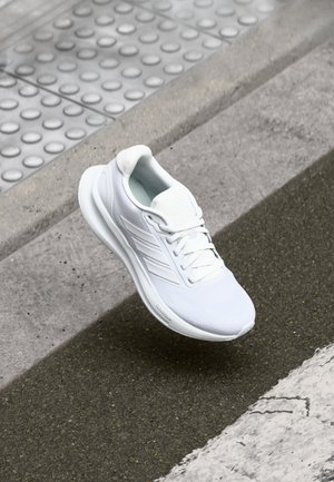 White athletic shoe with laces, floating above a textured sidewalk and asphalt road near a curb and tactile paving.