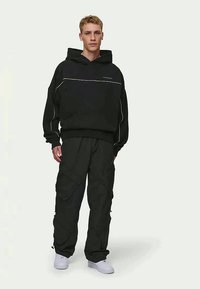 Sweatshirt preto com capuz e costura branca, combinado com calças cargo pretas largas com múltiplos bolsos e um corte descontraído. Ténis brancos.