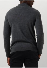 Man in een aansluitende, lange mouw grijze trui en zwarte broek, van achteren gezien met handen in de zakken.