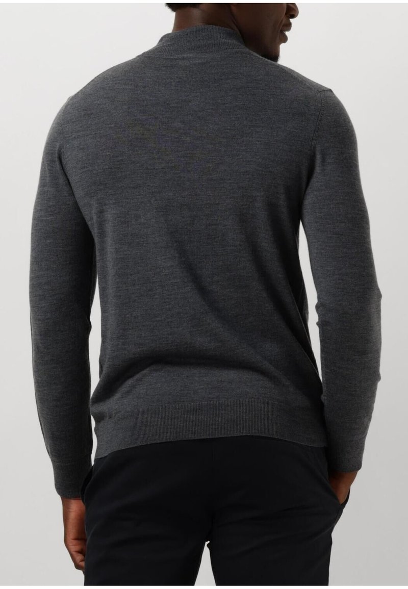 Mężczyzna w dopasowanym, długim szarym swetrze i czarnych spodniach, widoczny od tyłu z rękami w kieszeniach.