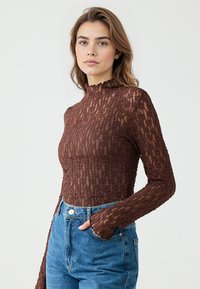 Bruine, gemêleerde top met lange mouwen en een hoge ruches-kragen, gecombineerd met hoge taille blauwe jeans. De stof heeft een golvend patroon.