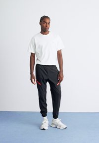 Mand med fletninger iført hvid t-shirt, sorte joggers med røde lynlåse og hvide sneakers, stående på lyseblå gulv mod hvid væg.