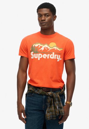 Mand iført en lys orange Superdry t-shirt med bjerg- og solgrafik, mørk ternet skjorte bundet om livet, blå jeans og armbåndsur.