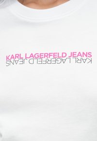 Valkoinen puuvillapaita, jossa on rohkealla pinkillä ja mustalla tekstillä "KARL LAGERFELD JEANS". Tasainen pinta ja pyöreäkauluksinen muotoilu.