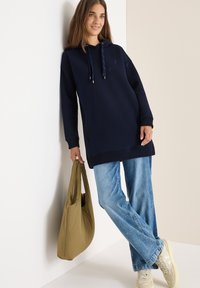 Felpa con cappuccio navy e cordoncini, tessuto testurizzato, maniche lunghe, abbinata a jeans azzurri e una borsa a tracolla color khaki.