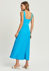 A blue pleated midi dress with a fitted bodice, wide straps, and an open back, made from a smooth, stretchy fabric.