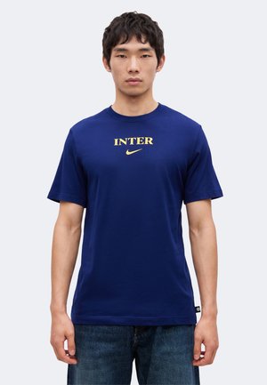 Άνδρας που φοράει μπλε μπλουζάκι με τη λέξη "INTER" και χρυσό λογότυπο Nike, συνδυασμένο με μπλε τζιν, στέκεται σε απλό ανοιχτό φόντο.