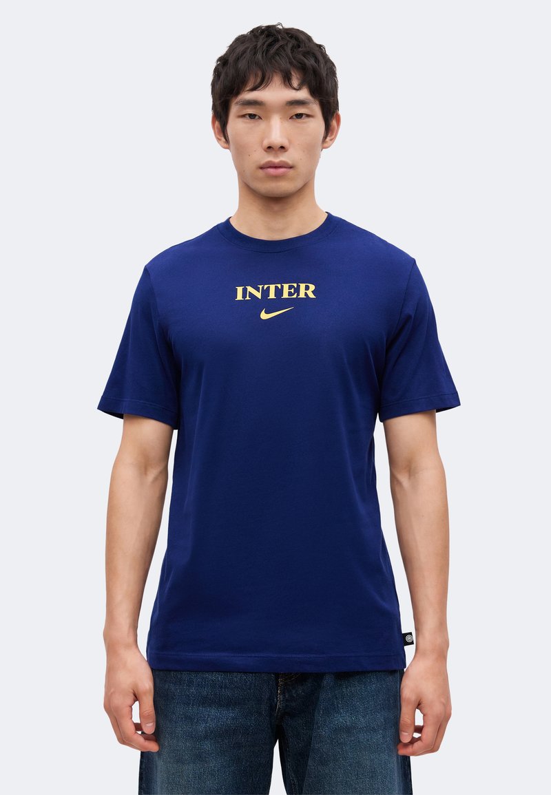 Άνδρας που φοράει μπλε μπλουζάκι με τη λέξη "INTER" και χρυσό λογότυπο Nike, συνδυασμένο με μπλε τζιν, στέκεται σε απλό ανοιχτό φόντο.