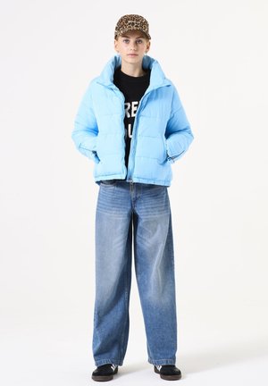 Veste en doudoune bleu clair avec col montant, fermeture éclair et deux poches latérales, associée à un jean en denim à jambes larges et une casquette à imprimé léopard.
