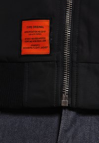 Zwarte bomberjack met een gebreide kraag en tailleband, voorzien van een zilveren rits en een oranje label met specificaties.