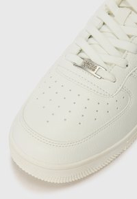White leather sneaker with perforated toe, flat laces, and a silver logo accent. Features textured rubber sole with a subtle pattern.