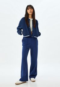 Marinträna blå idrottsjacka med vit kant och knappar, i kombination med matchande vida byxor. Slät tyg, avslappnad passform och avslappnade sneakers.
