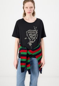 Zwart t-shirt met een glinsterend grafisch logo, gecombineerd met een gestreepte trui die om de taille is geknoopt in groen, rood en marineblauw. Lichte denim jeans.