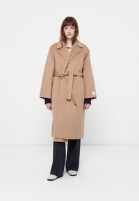 Cappotto a bellezza in misto lana beige con maniche larghe e colletto a punta, indossato sopra un outfit scuro, caratterizzato da un minimo dettaglio di branding.