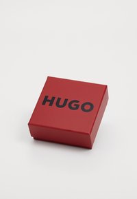 Röd, fyrkantig papperslåda med en framträdande svart "HUGO"-logotyp på locket. Slät yta med en ren, modern design.