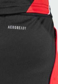 Sorte atletiske shorts med røde detaljer, med en glat tekstur og et "AEROREADY" logo i grå. Lynlåsdetalje synlig på siden.