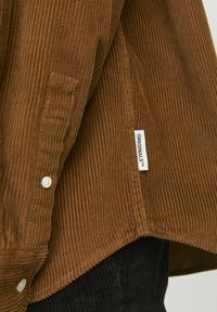 Chemise en velours côtelé marron avec une surface texturée, des boutons à l'avant, et une étiquette visible portant la mention "ORIGINALS" à l'ourlet.