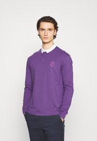 Mann mit den Händen in den Taschen, der ein lila Langarm-Polo-Shirt mit weißem Kragen und einem rosa heruntertropfenden Smiley-Gesicht-Logo auf der Brust trägt.