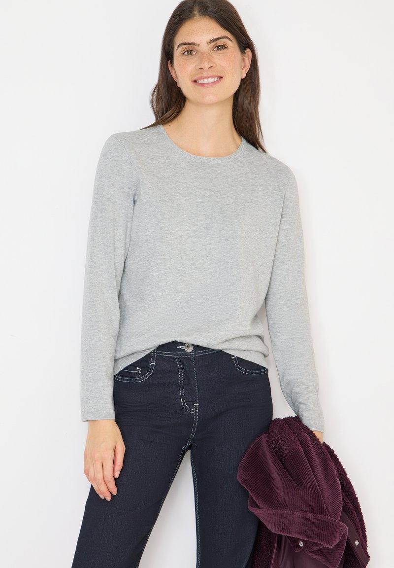 Maglietta grigia a maniche lunghe realizzata in tessuto morbido, indossata con jeans scuri. Un capo esterno color bordeaux è tenuto in una mano; presenta motivi e design minimi.