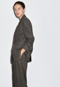 Giovane donna con i capelli raccolti, che indossa un blazer oversize grigio scuro e pantaloni abbinati, in piedi con una mano in tasca su uno sfondo bianco.