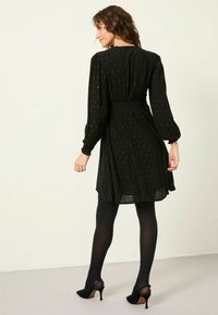 Schwarzes Kleid mit langen Ärmeln und einem gerafften Bund, das ein dezentes Muster aus goldenen Punkten aufweist. Kombiniert mit schwarzen Strumpfhosen und hohen Schuhen.