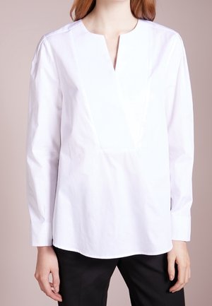 Femme portant un chemisier blanc à manches longues avec un col en encoche et un pantalon noir, debout devant un fond clair.