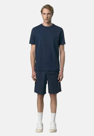 Giovane uomo in piedi, indossa una maglietta blu navy a maniche corte, pantaloncini abbinati, calzini bianchi e scarpe da ginnastica bianche, su uno sfondo chiaro e semplice.