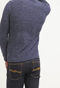 Pull en maille bleu marine avec une finition texturée, des poignets et un ourlet côtelés. Associé à un jean denim foncé avec des coutures jaunes en forme de vagues sur les poches.