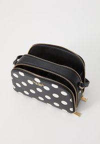 Sac à pois noir et blanc avec une texture lisse, double zips et accents en métal doré. Design rectangulaire compact avec deux compartiments.