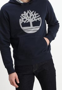 Sudadera azul marino con un gran logotipo de árbol gris en la parte delantera, bolsillo tipo canguro y capucha con cordones. Material suave y puños acanalados.