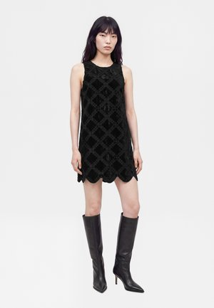 Une femme porte une mini-robe noire à motifs, sans manches et à l’ourlet festonné, ainsi que des bottes à talons montant jusqu’aux genoux, sur un fond blanc uni.