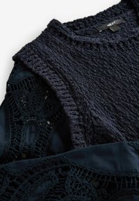 Donkerblauwe trui met een gestructureerde breiwerk, voorzien van een geribde halslijn en kantdetails op de mouwen, gecombineerd met een effen stof eronder.