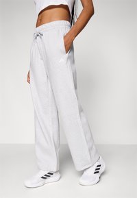 Calças de sweatshirt cinza claro com cintura elástica e cordão, apresentando bolsos laterais e um logótipo da Adidas na parte superior da coxa esquerda.