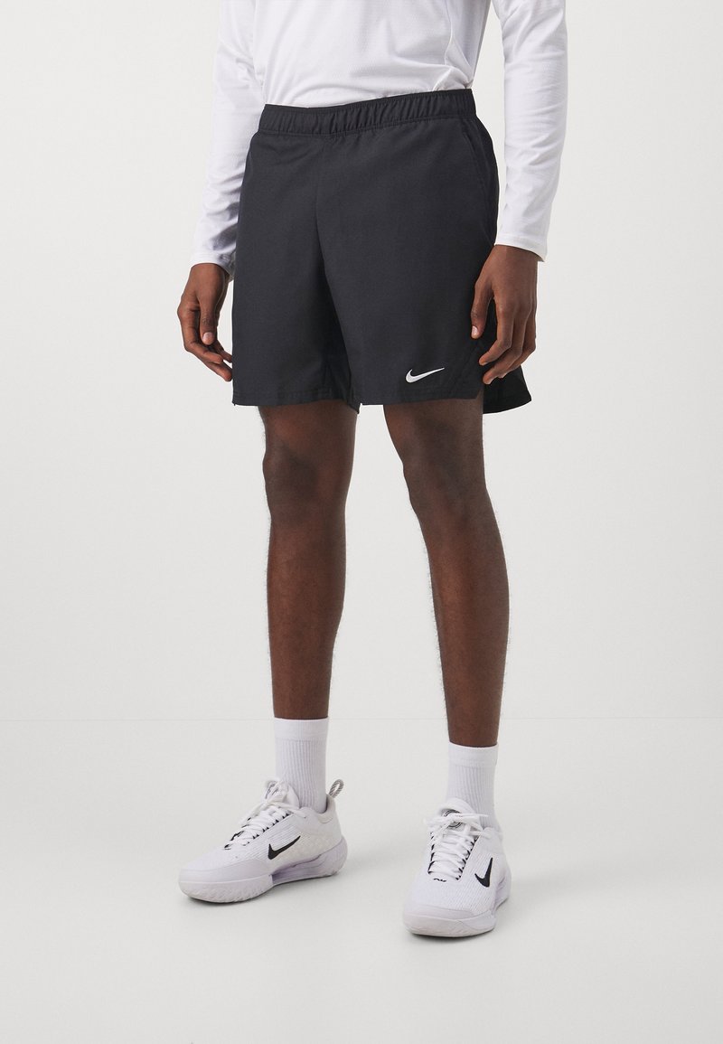 Homme portant un short de sport noir Nike, une chemise blanche à manches longues, des chaussettes blanches et des baskets Nike blanches, debout sur un fond uni.