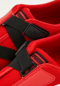 Røde idrettssko med nettingpaneler og gummidetaljer, med svarte stropper som har Puma-logo. Rund tå og polstret innside.
