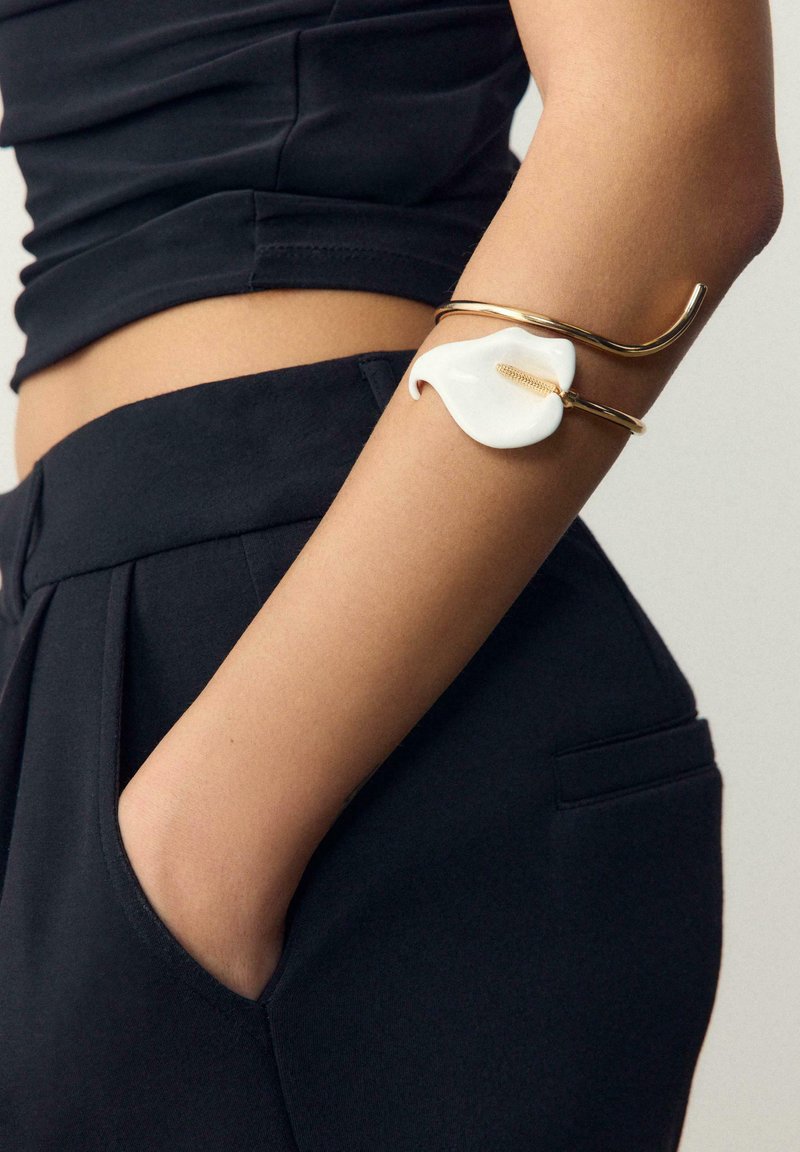 Bras orné d'un bracelet en or avec un motif de calla blanc, reposant près de la poche d'un pantalon noir taille haute et d'un haut noir.