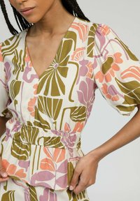 Blouse à motif floral en crème avec des accents verts, roses et orange. Présente un col en V, des manches courtes puff et une taille élastique.