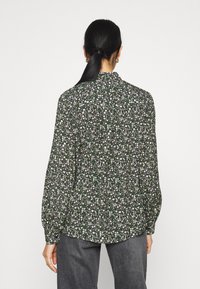 Grüne Blumenbluse mit langen Ärmeln, lässiger Passform und geknöpftem Rücken. Sie zeigt kleine weiße und gelbe Blumen auf einem dunklen Hintergrund.