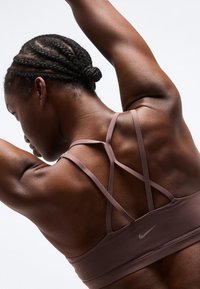 Močna oseba s kitkami in raztegnjenimi rokami nad glavo, oblečena v sivordečo športno modrček znamke Nike s križanimi trakovi na belem ozadju.