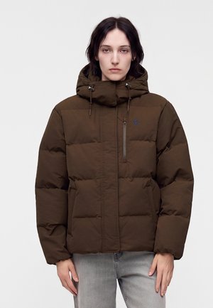 Jeune personne aux cheveux foncés portant une veste matelassée marron avec une capuche et un jean gris clair, debout devant un fond blanc uni.