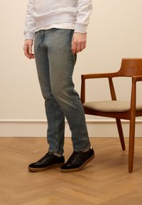 Jeans in denim blu con taglio dritto, indossati con sneakers nere. Lo sfondo presenta una sedia di legno e una parete di un colore chiaro.