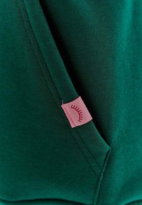 Zelená bavlněná tkanina s hladkým povrchem, s růžovou etiketou s grafikou oka zašitou na bočním švu.