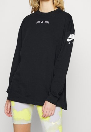 Frau trägt ein schwarzes langärmliges Nike Air Shirt und gelb-graue Nike-Shorts, steht mit entspannten Händen an den Seiten.