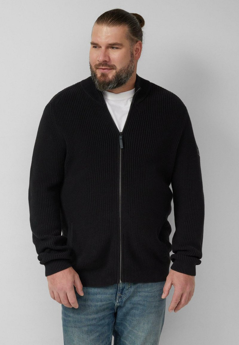 Zwarte geribbelde zip-up trui met lange mouwen, een hoge kraag en zijzakken. Gedragen over een wit t-shirt, in combinatie met spijkerbroek.