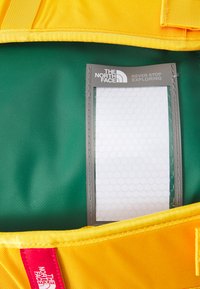 Yellow fabric backpack with a green interior, featuring a gray label holder and hexagonal pattern on the label. Red logo tag visible.