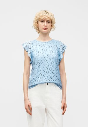 Νεαρό άτομο με κοντά, σγουρά ξανθά μαλλιά, φορούν ένα ανοιχτό μπλε μπλουζάκι με κοντά μανίκια και λευκό παντελόνι, στέκεται μπροστά σε ένα απλό φόντο.