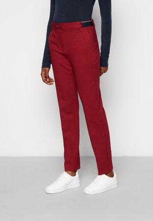 Persoon die nauwsluitende rode broek, donkerblauw shirt met lange mouwen en witte sneakers draagt, staand tegen een effen lichte achtergrond.